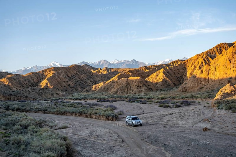 Off-road vehicle driving through a canyon, landscape of eroded hills at sunrise, badlands, white mountain peaks of the Tian Shan Mountains in the background, Canyon of the Forgotten Rivers, Issyk Kul, Kyrgyzstan