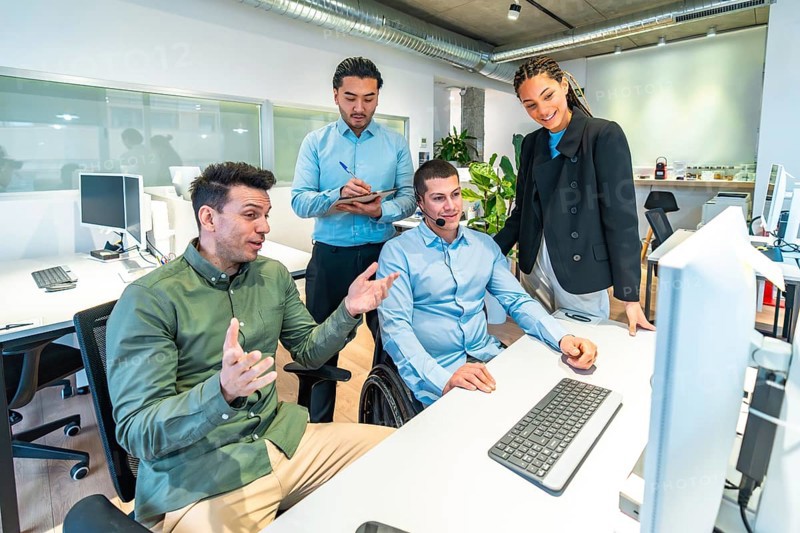 Diverse business team collaborating on a computer project, demonstrating inclusion and teamwork in the workplace with a disabled colleague in a wheelchair