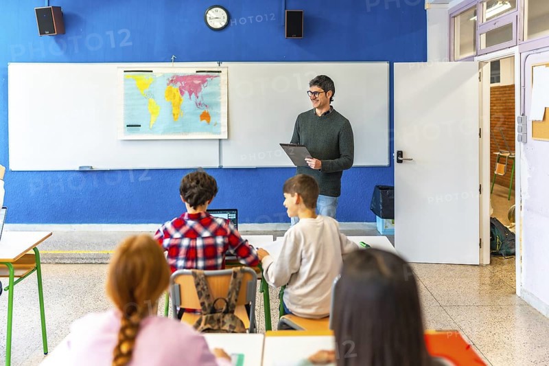 Geography teacher explaining a lesson holding a clipboard and smiling to elementary school students in a classroom