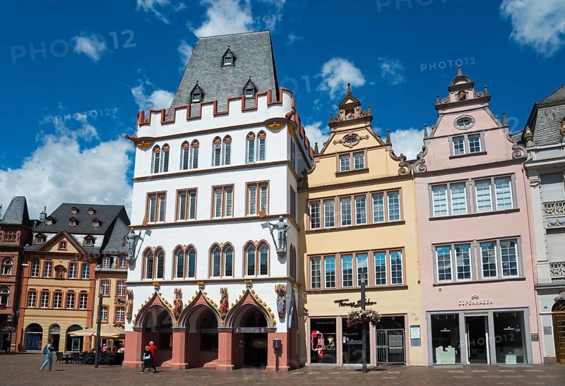 Colourful historic buildings in Gothic style with towers and windows on a lively town square under a blue sky, on the left Haus Steipe with arcade and battlements, Hauptmarkt, Trier, Rhineland-Palatinate, Germany