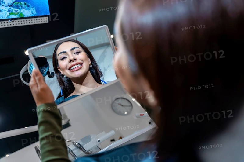 Patient looking at her whiter teeth in the mirror at the dental clinic, satisfied with the results of the cosmetic dental treatment