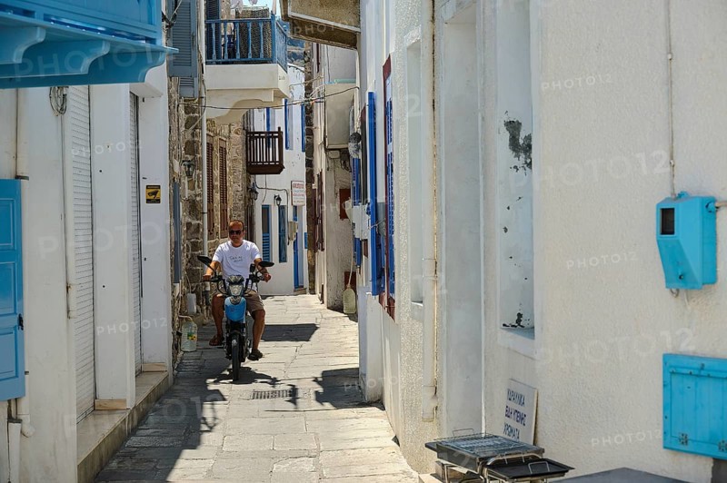A narrow white alley with blue shutters. A man rides a motorbike through the sun, Mandraki, Nisyros, Dodecanese, Greek Islands, Greece