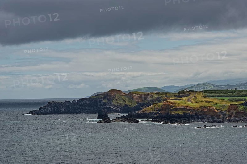 A rocky coastline with green hills in the background under a cloudy sky, Fenais da Luz, Sao Miguel Island, Azores, Portugal