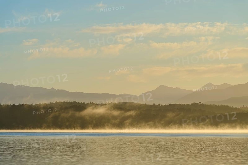 Fog over a calm lake under a clear sky with mountains in the background, Lake Mahinapua, Ruatapu, South Island, New Zealand