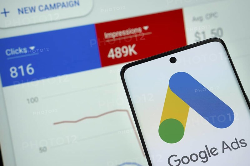 Google Ads logo is displayed on a smartphone screen, with a stock graph webpage visible on a computer screen in the background