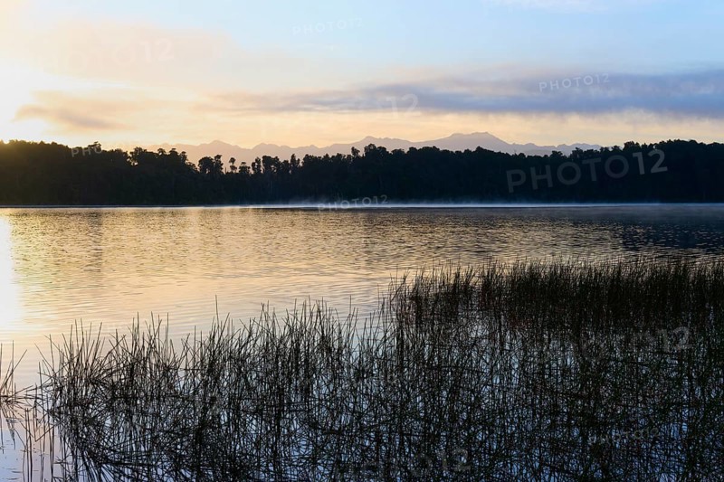 Lake in front of rising sun with plants and mountain silhouette, Lake Mahinapua, Ruatapu, South Island, New Zealand