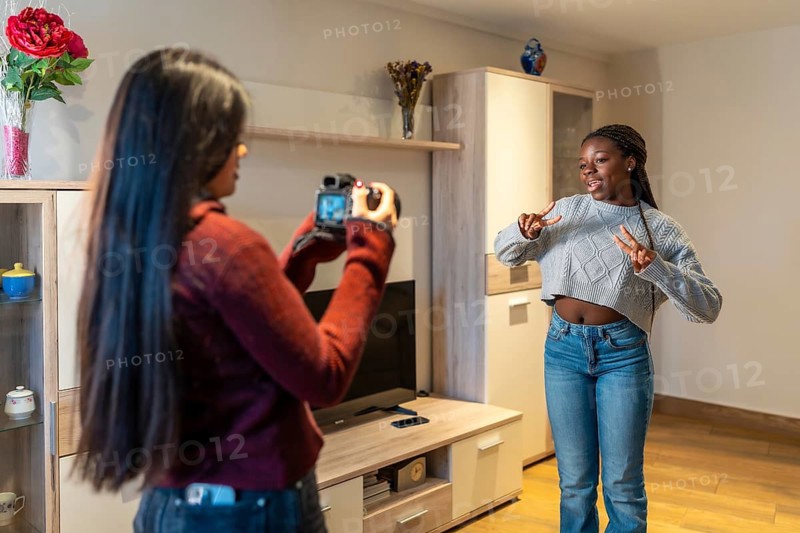 Two teenage girls are having fun at home, one filming the other dancing with a photo camera