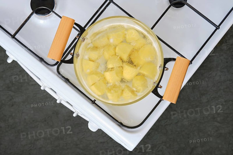 Sliced yellow potatoes are boiling in a clear glass pot on a white stove with wooden handles. The kitchen has a modern design, highlighting the process of preparing a meal