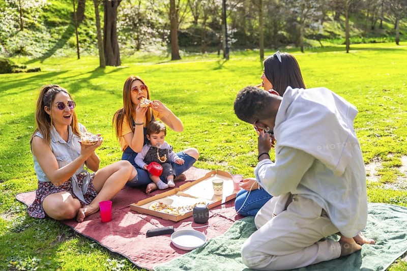 Group of friends savoring delicious pizza while enjoying a sunny picnic in a vibrant park, surrounded by laughter and good company