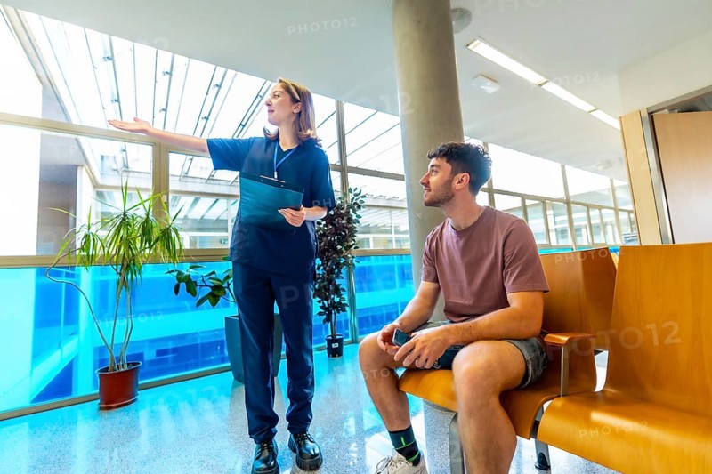 Female nurse holding a clipboard, providing instructions to a male patient seated in a bright, modern waiting room of a healthcare facility