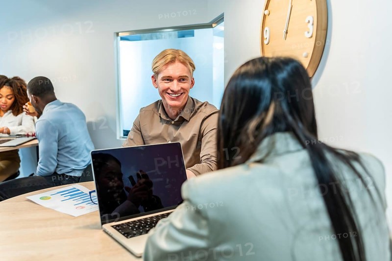 Two managers are smiling and talking during a meeting in a coworking office, using a laptop and analyzing graphs