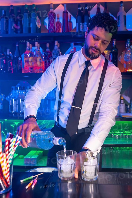 Professional bartender preparing cocktails, pouring alcoholic beverage into glasses with ice at bar counter in nightclub