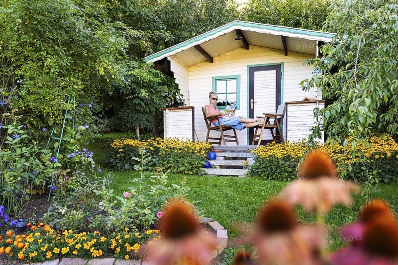 A woman sits relaxed on the small veranda of a garden shed and reads. Blooming summer flowers in the garden. Germany