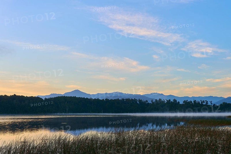 A calm, misty lake in morning sunlight with mountains in the background, Lake Mahinapua, Ruatapu, South Island, New Zealand