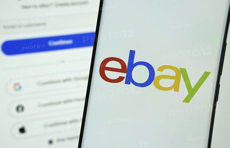 ebay logo is displayed on a smartphone screen, with the ebay login page visible on a computer screen in the background