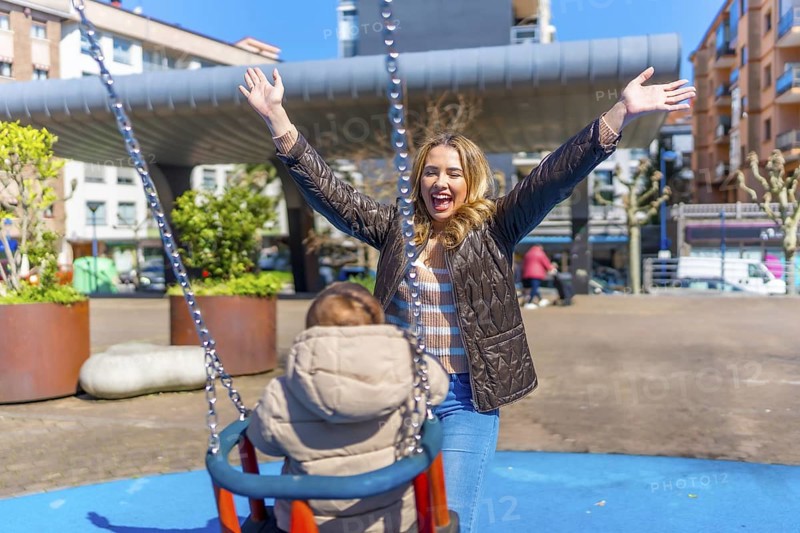 Young mother having fun pushing her baby on a swing in an urban playground, enjoying a sunny day outdoors