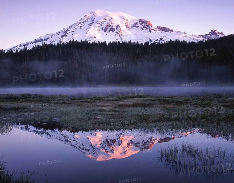 Snow capped mountain reflecting in a calm lake at dusk, surrounded by mist and lush forest. The sun graces the peak, creating an enchanting and serene landscape