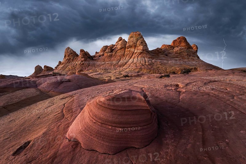 A breathtaking view of Coyote Buttes in Paria Canyon-Vermilion Cliffs Wilderness, Arizona, under stormy skies. The dramatic clouds and a striking lightning bolt accentuate the unique red rock structures