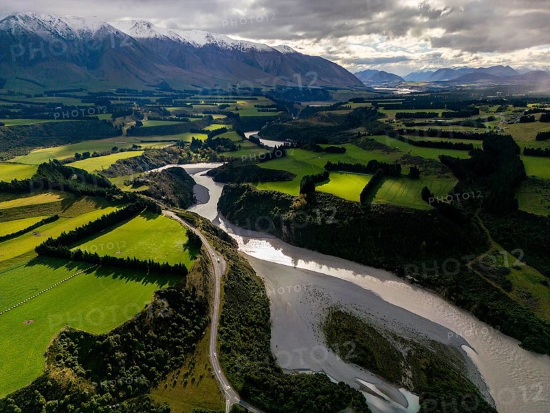 Aerial view of Canterbury region's lush green farmland and winding river, nestled against the backdrop of snow-capped mountains, capturing the essence of New Zealand's natural beauty