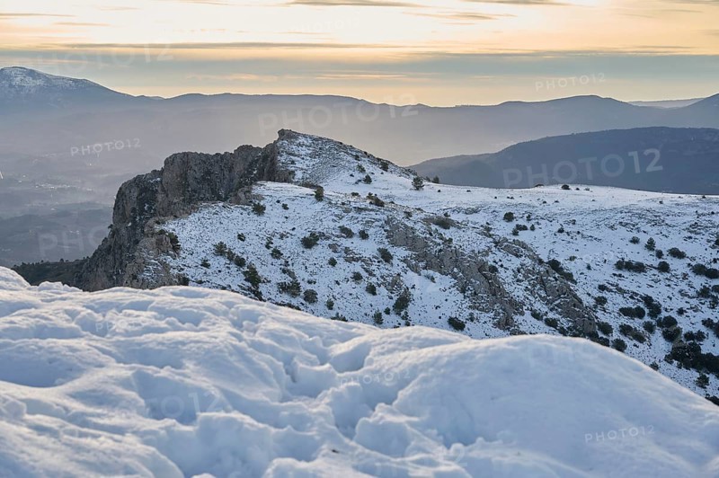 Scenic view of Montcabrer, Spain, showcasing its snow-covered peaks under a softly glowing sunrise. The tranquil landscape captures the serene beauty of winter