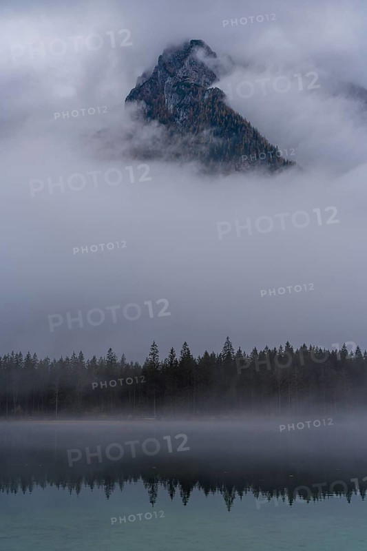 A stunning view of a mountain peak in Hintersee, partially shrouded in fog, surrounded by serene forest and reflected in a tranquil lake, capturing the ethereal beauty of the Bavarian Alps