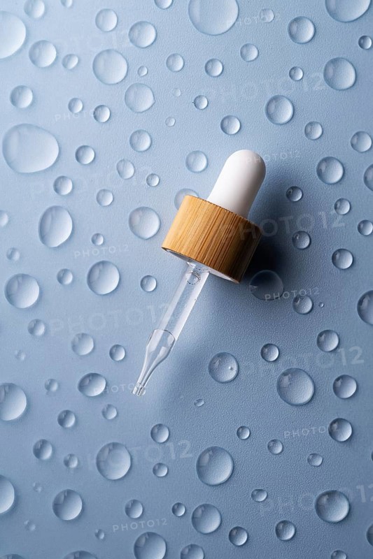 A glass pipette bottle lies on a textured blue surface Water droplets surround the bottle, emphasizing freshness and hydration in skincare