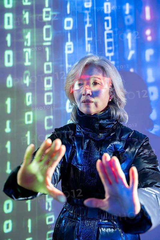 An elderly Asian woman engages with the metaverse using virtual reality technology. She wears futuristic glasses, immersed in a digital environment with binary code projections