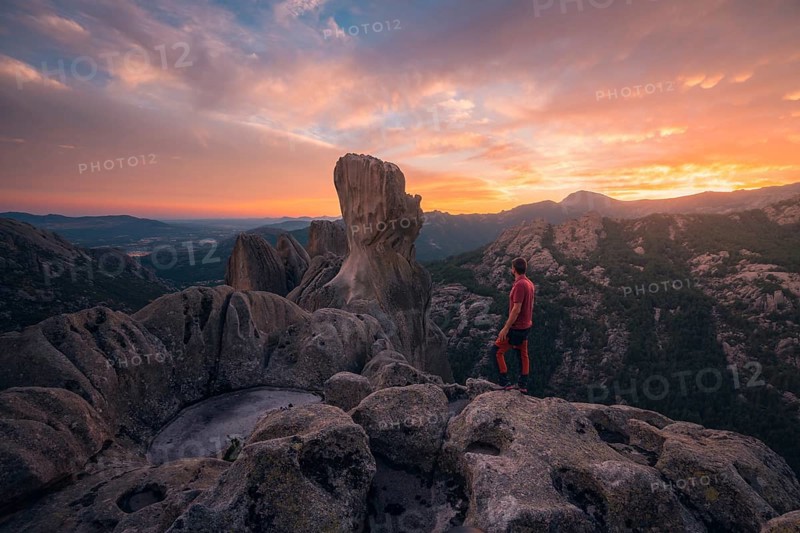 Back view of unrecognizable man standing on rocky outcrop, gazing at vibrant sunset over Pedriza Caballeros. Majestic rock formations and expansive views of Sierra de Guadarrama