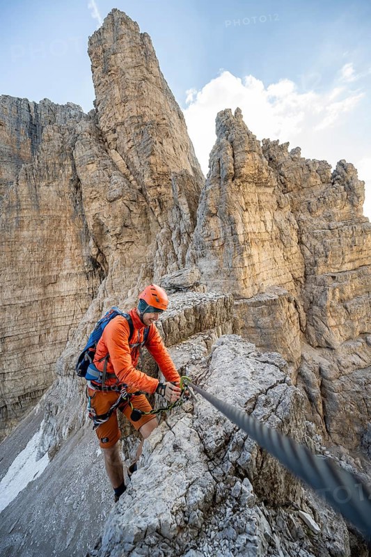Mountaineer climbs an exposed rock in the secured Via Ferrata Bocciere Centrale via ferrata, spectacular rock towers in the back, Brenta Mountains, Brenta-Adamello Natural Park, Trentino, Italy