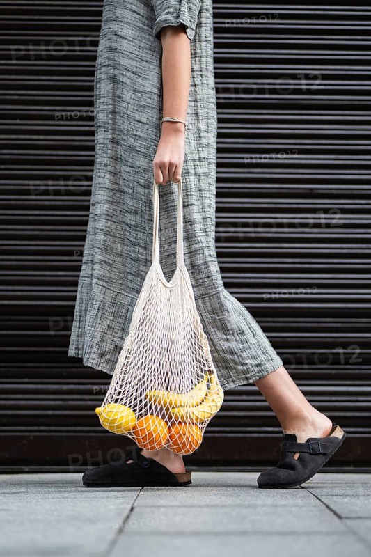 A woman in a long, gray dress carries a reusable mesh bag filled with bananas and oranges. The individual walks on a sidewalk, wearing black slip-on shoes
