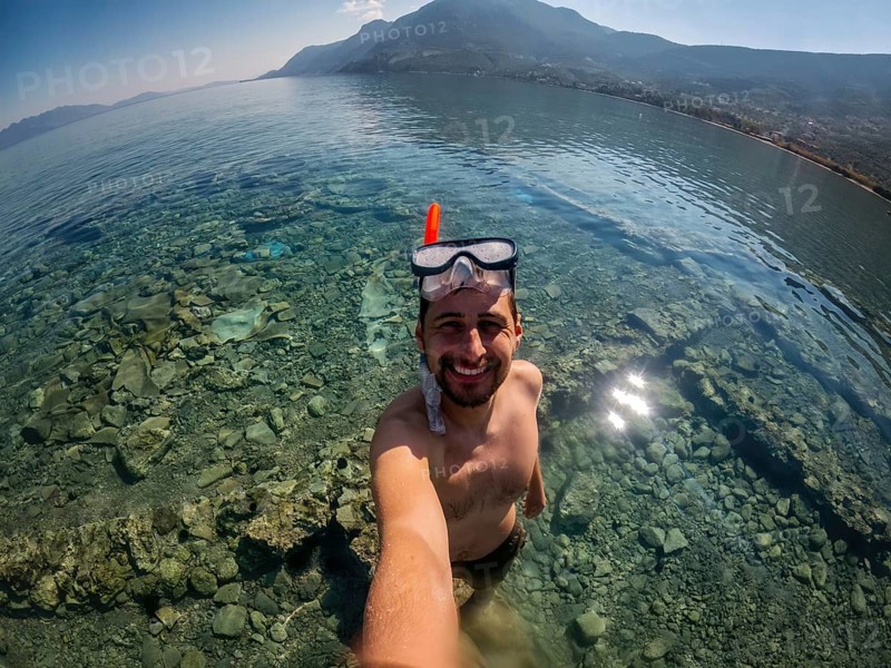 A man enjoys snorkeling in Greece, exploring the crystal-clear waters and rocky seabed. The scenic landscape with mountains and bright sky adds to the serene experience