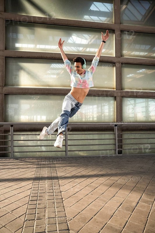 A joyful dancer in a colorful cropped top and jeans leaps gracefully, wearing headphones and sneakers. The urban industrial setting enhances the vibrant energy and motion