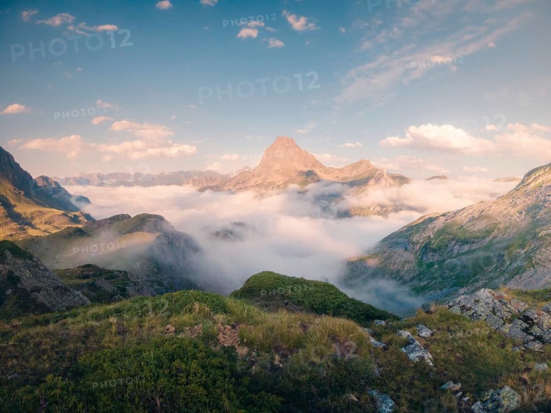 Serene mountain peak rises above a lush green landscape enveloped in wispy clouds under a vast blue sky in summer. The scene captures the essence of tranquility and natural beauty in the Pyrenees