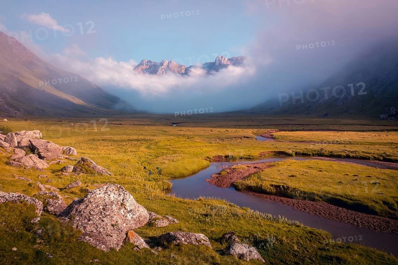 A serene mountain valley landscape with a winding river and mist shrouded peaks in summer. The warm sunlight gently illuminates the lush green terrain and rocky foreground in the Pyrenees