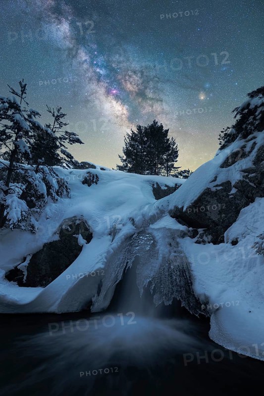 Captivating night scene depicting the Milky Way over a snow-laden landscape in Iceland A frozen waterfall adds an enchanting touch to this serene winter wonderland