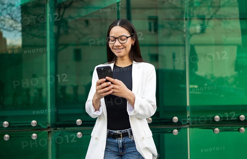 Hispanic businesswoman wearing glasses and a white blazer, smiling while using her smartphone against a modern glass building background. Urban business lifestyle scene