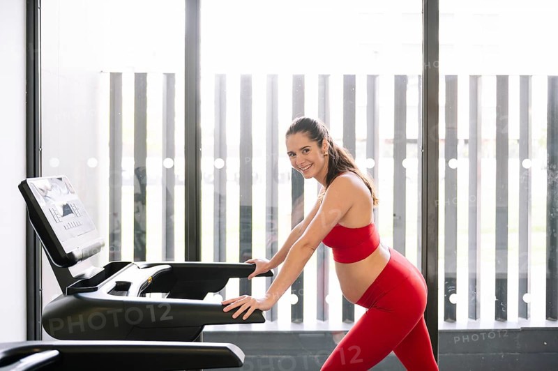 Pregnant woman in a red sports outfit exercising on a treadmill, promoting pregnancy wellness and active lifestyle. Bright, modern gym environment with large windows