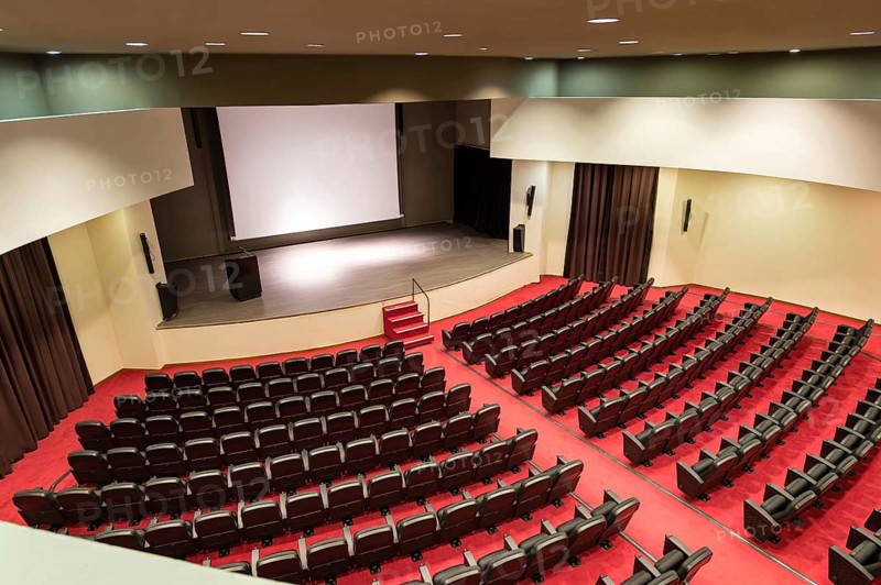 An empty convention center featuring rows of seats facing a wooden stage and a large white screen, ideal for presentations or performances, under soft lighting