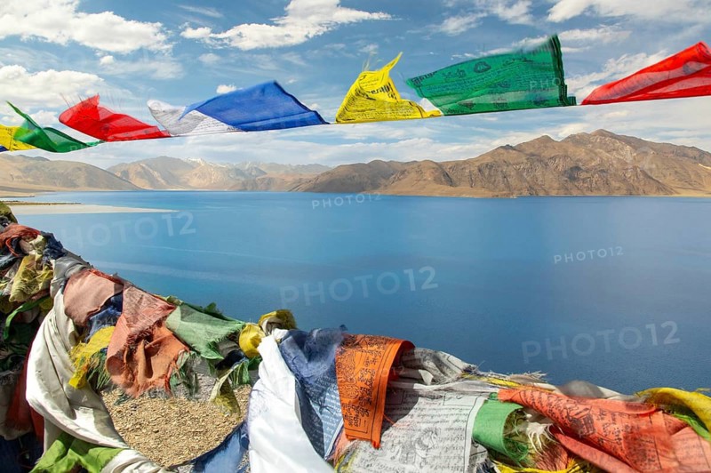 Colorful prayer flags flutter against the backdrop of the serene Pangong Tso lake, set amidst the rugged mountains of Ladakh, India, under a vibrant blue sky