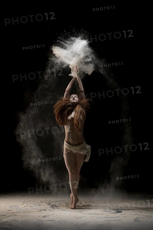 Slim young barefoot woman throwing sand over head while dancing against black background