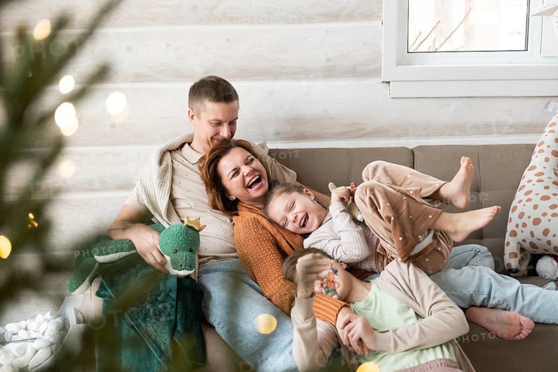 A joyful family celebrates Christmas in a cozy home setting. Two adults and two children, all laughing and cuddling on a sofa, are surrounded by Christmas decorations and soft lighting