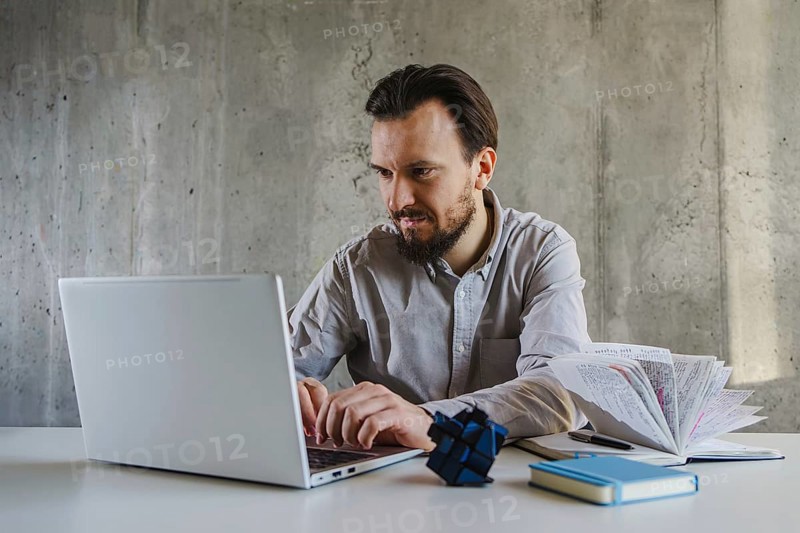 A man in a shirt works intently on a laptop at office. Beside him are a book and a Rubik cube, suggesting multitasking or problem solving in a business setting