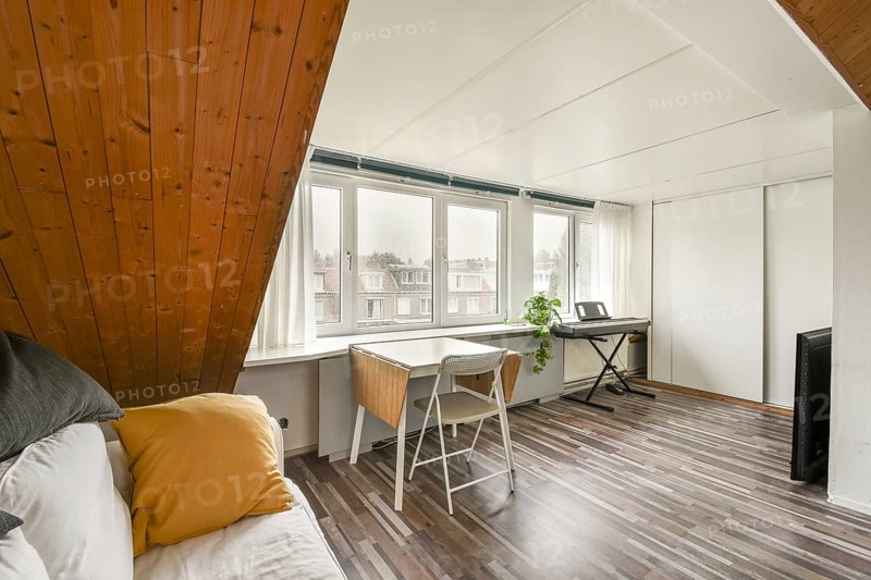 Spacious attic room featuring a slanted wooden ceiling, bright windows with a view, a cozy workspace with a white desk, and a keyboard, creating a perfect creative setting