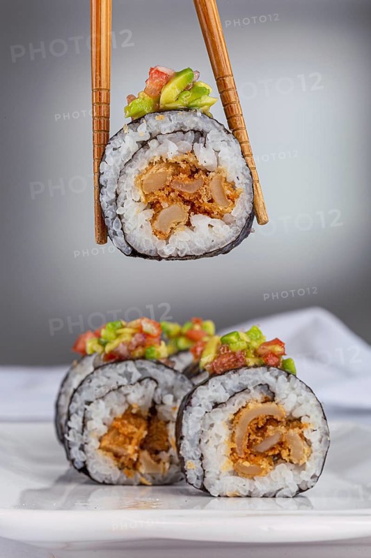 A close up of a sushi roll with vibrant avocado and salmon topping, held by chopsticks with more rolls above a white background. Perfect for food and restaurant themes
