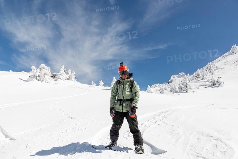 A male snowboarder stands confidently in a pristine snowy landscape under a clear blue sky, wearing a green jacket and orange helmet