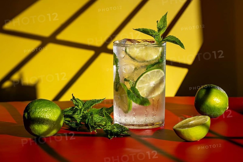 A refreshing classic mojito with lime and mint is set against a vibrant yellow and yellow backdrop. The cocktail features fresh lime slices and mint, placed on a smooth red surface