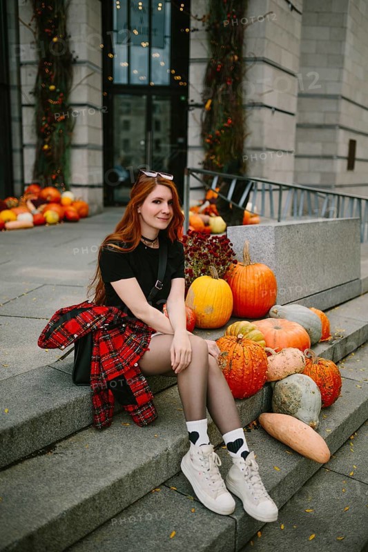 A young woman in autumnal attire sits on stone steps adorned with pumpkins in an urban Montreal setting, capturing the essence of fall with vibrant seasonal decor