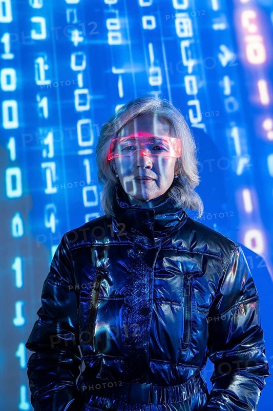 An elderly Asian woman wearing futuristic eyewear is immersed in the metaverse. Blue digital code surrounds her, symbolizing virtual reality and technological advancement