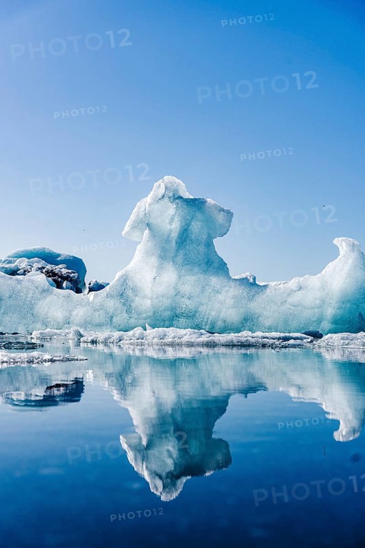 A majestic ice formation towers over the serene waters of Glacier Jokulsarlon in Iceland, reflecting its intricate shapes in the clear, still lagoon under a bright blue sky