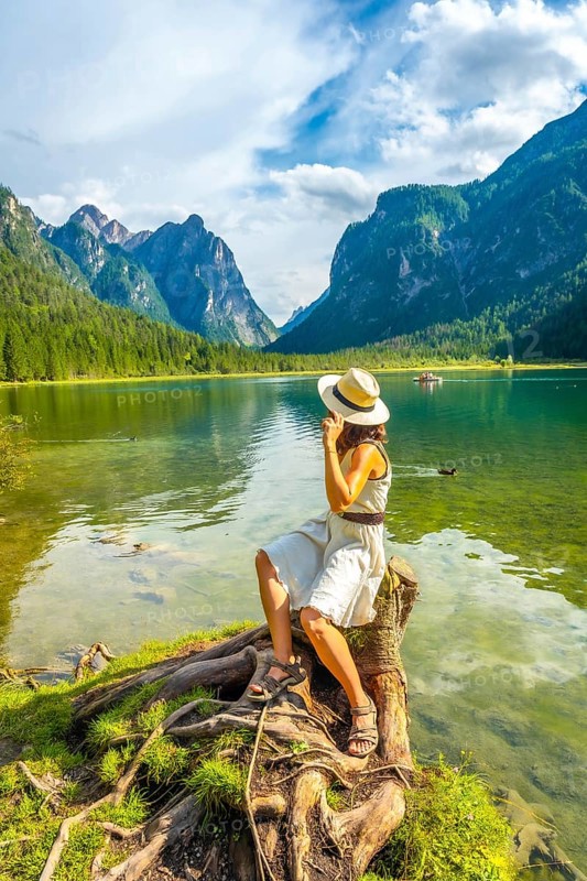Young woman wearing a straw hat sitting on a tree stump by lake dobbiaco, enjoying the breathtaking view of the dolomites on a summer day